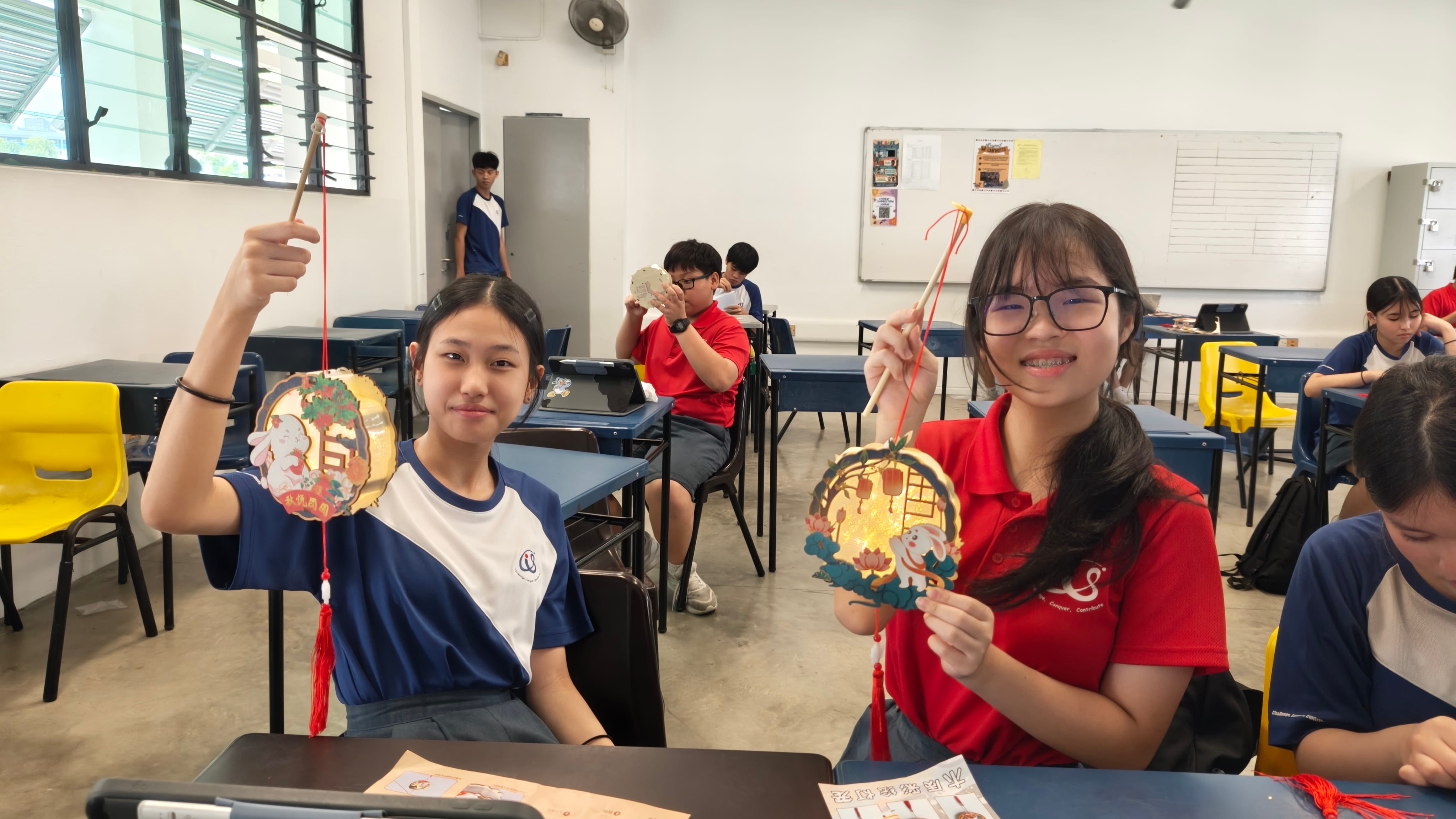 Two secondary school girls sit at their desks in a classroom, smiling and holding up their completed Mid-Autumn Festival lanterns featuring rabbit and lotus designs with the Chinese text 秋情圆圆.